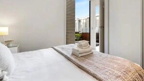 A modern hotel room with a neatly made bed, a small bedside table with a lamp, and a view of the outside balcony through a sliding door. - Lovely And Cozy Apartment - Parking, Pool And Gym! (Auckland)