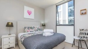 A modern bedroom with a large bed, a nightstand, a chair, and a window with a city view. - Wynyard Viaduct Harbour 2BR+Parking: Zodiak Stays (Auckland)
