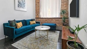 Una sala de estar con un sofá azul, una mesa de centro redonda, una araña, un ventanal con cortinas y un televisor montado en la pared.