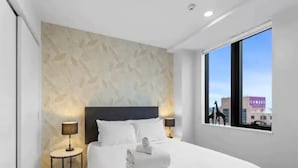 A modern hotel room with a large bed, two bedside tables, and a view of the city through a large window. - Stunning 2BR w/ Views Near Sky Tower- Zodiak Stays (Auckland)