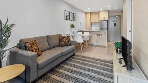 A modern living room with a grey sofa, a small wooden table, and a TV on a stand. A kitchen with a fridge and a dining area is visible in the background. - Stunning 2BR w/ Views Near Sky Tower- Zodiak Stays (Auckland)