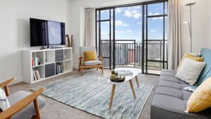 A modern living room with a flat-screen TV, bookshelves, a sofa, and a balcony with a city view. - Bright 1 Bedroom Beauty with Balcony Zodiak Stays (Auckland)