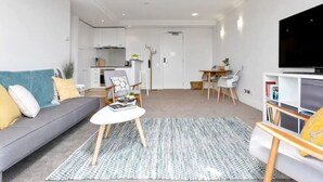 A modern living room with a grey sofa, a small white coffee table, and a TV on the right. There is a kitchen in the background with white cabinets and a dining area with a table and chairs. - Bright 1 Bedroom Beauty with Balcony Zodiak Stays (Auckland)