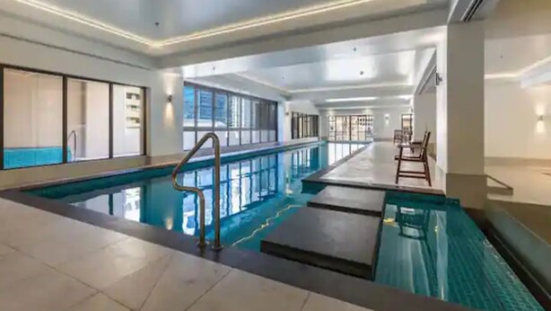 A large indoor swimming pool with a tiled floor, a metal ladder, and a long, narrow poolside area. - Bright 1 Bedroom Beauty with Balcony Zodiak Stays (Auckland)