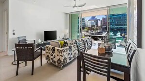 A modern living room with a sofa, dining table, and chairs. There is a TV and a ceiling fan. The room has large windows with a view of buildings and a body of water. - Gorgeous 2BR in Wynyard Quarter by Zodiak Stays (Auckland)