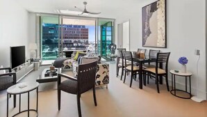 A spacious living area with a modern sofa, dining table, and chairs, large windows with a city view, and a ceiling fan. - Gorgeous 2BR in Wynyard Quarter by Zodiak Stays (Auckland)