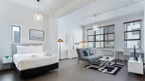 A modern hotel room with a large bed, a grey sofa, and a small table. There are two floor lamps and a large window with blinds. - Stylish studio near Spark Arena I Zodiak Stays (Auckland)