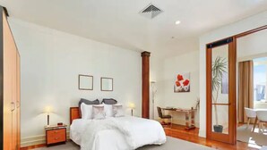 Une chambre spacieuse avec un grand lit, un bureau et une chaise. Il y a une armoire sur la gauche et une porte coulissante qui donne sur un balcon.