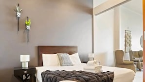 A modern hotel room with a large bed, a nightstand with a lamp, two wall-mounted plants, and a view of the outside through a large window.