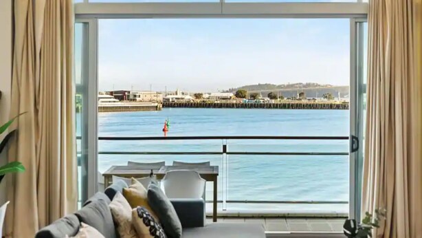 A room with a large window overlooking a waterfront, featuring a sofa and a table.