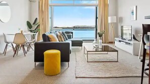 A modern living room with a sofa, ottoman, and coffee table, a dining area with chairs, and a large window offering a view of the sea.