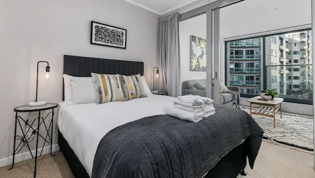 A modern hotel bedroom with a large bed, a view of the city, and a balcony with a view of the buildings. - Beautifully Modern Brand New Central Apartment (Auckland)