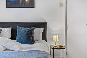 Un lit bien fait, avec une couette bleue, des oreillers blancs et une tête de lit noire. On y trouve une petite table de chevet avec une lampe et une boîte étiquetée « avec ».