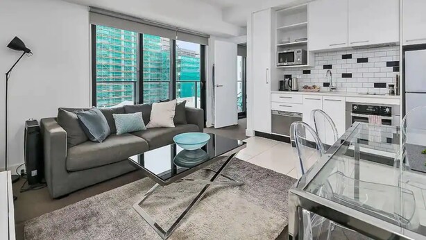 Een modern appartement met een grijze bank, een glazen salontafel en een keuken met witte kasten en apparatuur.