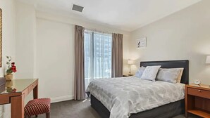 A hotel room with a bed, a desk, a chair, a nightstand, a lamp, and a window with curtains. - Pool, Gym And Style In The Cbd (Auckland)