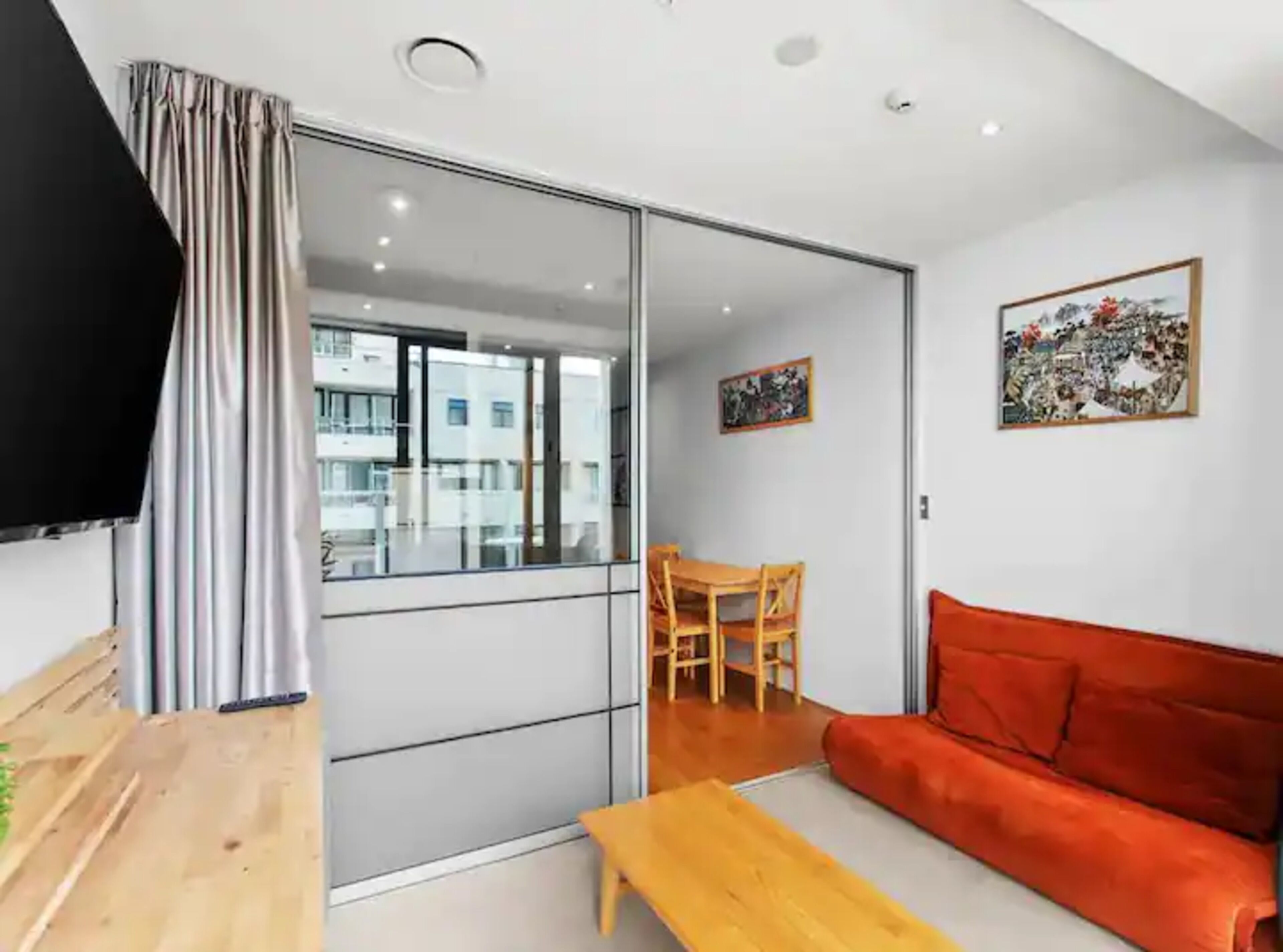 Ein modernes Wohnzimmer mit einer Schiebetür zum Balkon, einem Holztisch, einem roten Sofa und zwei gerahmten Kunstwerken an der Wand.