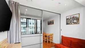 A modern living room with a sliding glass door leading to an outdoor balcony, a wooden table, a red sofa, and two framed artworks on the wall. - Stylish Central City 1-Bedroom With Patio (Auckland)