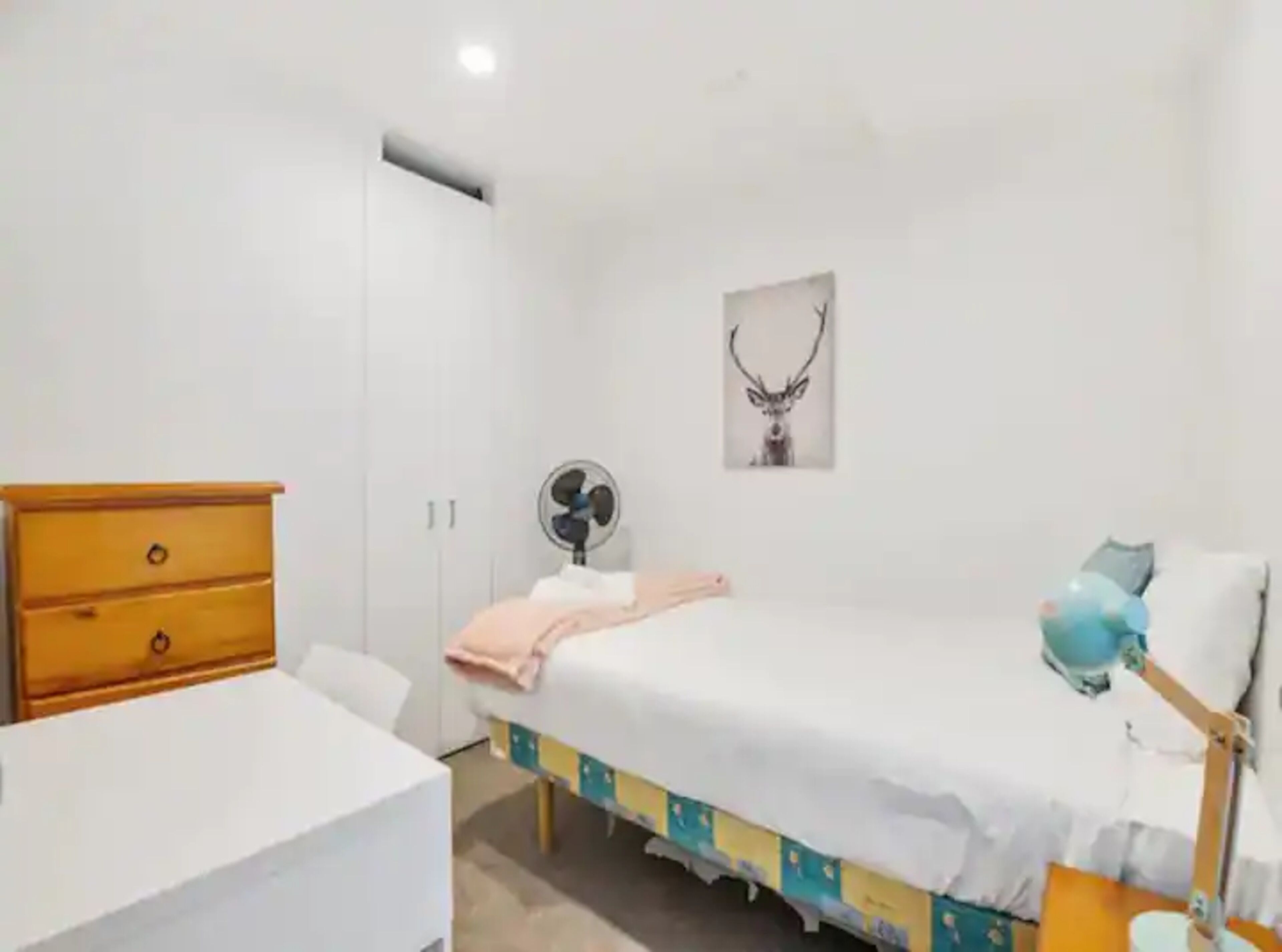 A bedroom with a bed, bedside table, dresser, and a wall-mounted fan.
