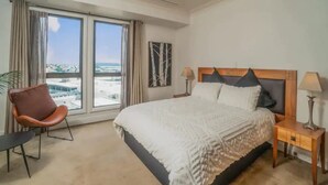 Ett hotellrum med en stor säng, en stol, ett nattduksbord och utsikt över staden genom stora fönster.