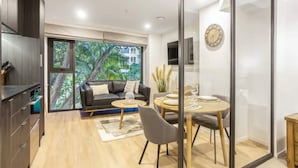 A modern living room with a sofa, coffee table, and dining area. There is a large window with a view of greenery and a building. - Condo in Queen St CBD w Pool and Gym Zodiak Stays (Auckland)