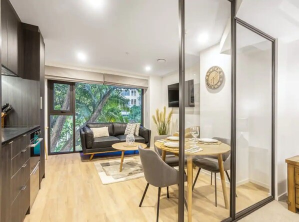 A modern living room with a sofa, coffee table, and dining area. There is a large window with a view of greenery and a building. - Condo in Queen St CBD w Pool and Gym Zodiak Stays (Auckland)