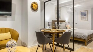 A modern room with a bed, a dining table with chairs, a TV mounted on the wall, and a decorative clock. - Condo in Queen St CBD w Pool and Gym Zodiak Stays (Auckland)