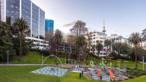 A park with playground equipment, palm trees, and modern buildings in the background. - Condo in Queen St CBD w Pool and Gym Zodiak Stays (Auckland)