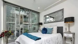 Een moderne slaapkamer met een groot bed, nachtkastje, spiegel en een raam met jaloezieën.