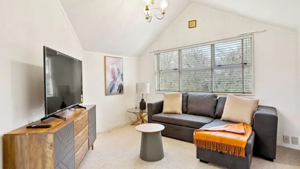 A modern living room with a flat-screen TV, a wooden cabinet, a grey sofa, and a small round table. - Charming 3 Bedroom Home In Tranquil Rural Karaka (Karaka)