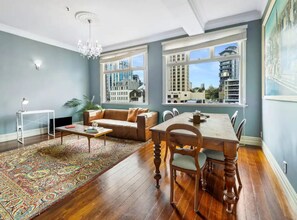 Sala de estar com sofá, mesa de jantar, cadeiras, lustre e uma grande janela com vista para a cidade.