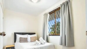 A neatly made bed with white linens and patterned pillows, a small bedside table, and a window with grey curtains.