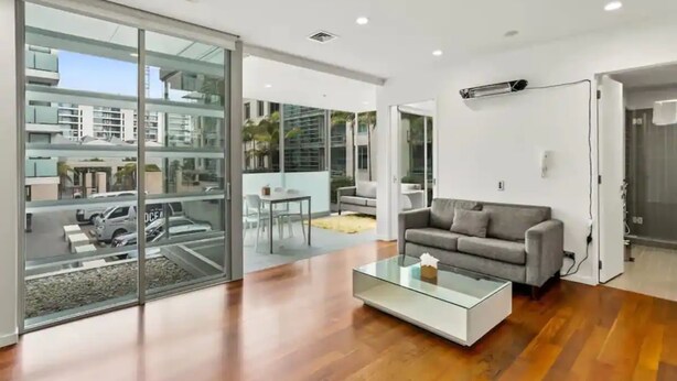 Uma sala de estar moderna com uma mesa de centro de vidro, um sofá cinzento e vista para a rua e edifícios através de grandes portas de vidro.