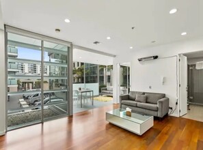 Ein modernes Wohnzimmer mit einem Glastisch, einem grauen Sofa und Blick auf eine Straße sowie Gebäude durch große Glasüren.
