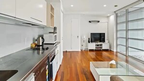 Cozinha moderna com exaustor de aço inoxidável, micro-ondas e chaleira. Piso de madeira e mesa de vidro com um vaso. Uma TV está montada na parede ao fundo.