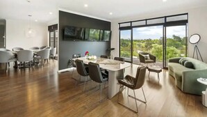 A modern living space with a dining area, kitchen, and a large sliding glass door leading to a balcony with a view of greenery. - North Shore Mansion With 20M Pool (Auckland)