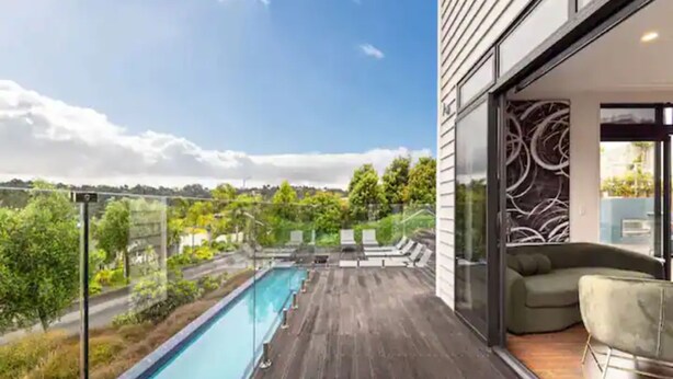 A modern outdoor pool area with a wooden deck, lounge chairs, and a glass railing overlooking a lush landscape. - North Shore Mansion With 20M Pool (Auckland)