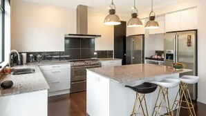 A modern kitchen with a large island, stainless steel appliances, pendant lighting, and a bar area with stools. - North Shore Mansion With 20M Pool (Auckland)