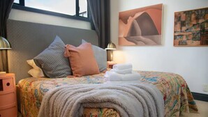 A neatly made bed with a floral bedspread, multiple pillows, and a blanket. There are two bedside tables with lamps, a window with curtains, and a painting on the wall. - One-bedroom loft in Viaduct by Zodiak Stays (Auckland)