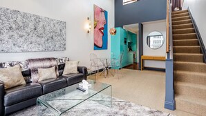 A modern living room with a black leather sofa, a glass coffee table, and a large abstract painting. There is a staircase leading to an upper level with a view of the city. - One-bedroom loft in Viaduct by Zodiak Stays (Auckland)