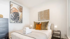 Un dormitorio con una cama, una mesita de noche con lámpara, un aparador y dos obras de arte en la pared.