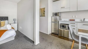 A modern hotel room with a bed, a bedside table with a lamp, a small desk with a chair, a kitchenette with a fridge, microwave, and oven, and a large window. - Tidy Apartment With Balcony (Auckland)