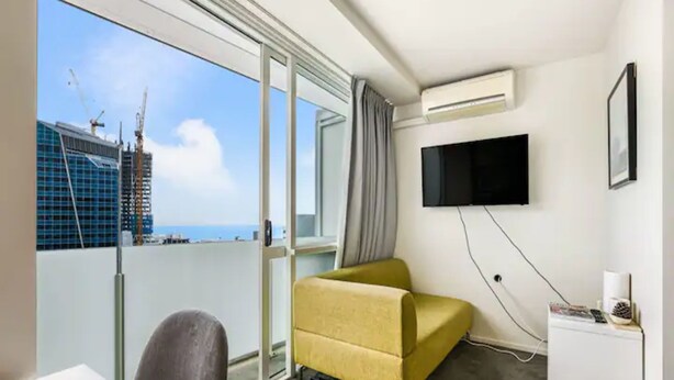 A hotel room with a large sliding glass door leading to a balcony, a flat-screen TV mounted on the wall, a small white desk, and a yellow sofa. - Tidy Apartment With Balcony (Auckland)