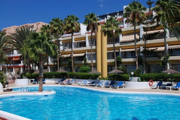 A swimming pool with lounge chairs and palm trees, surrounded by a multi-story hotel building.