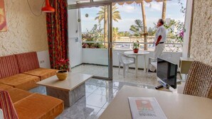 Un salon moderne avec un canapé, une table basse et un téléviseur. Il y a un balcon avec vue sur des palmiers et une piscine.