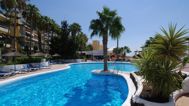 Une piscine entourée de bâtiments d’hôtel, avec des chaises longues et des palmiers.