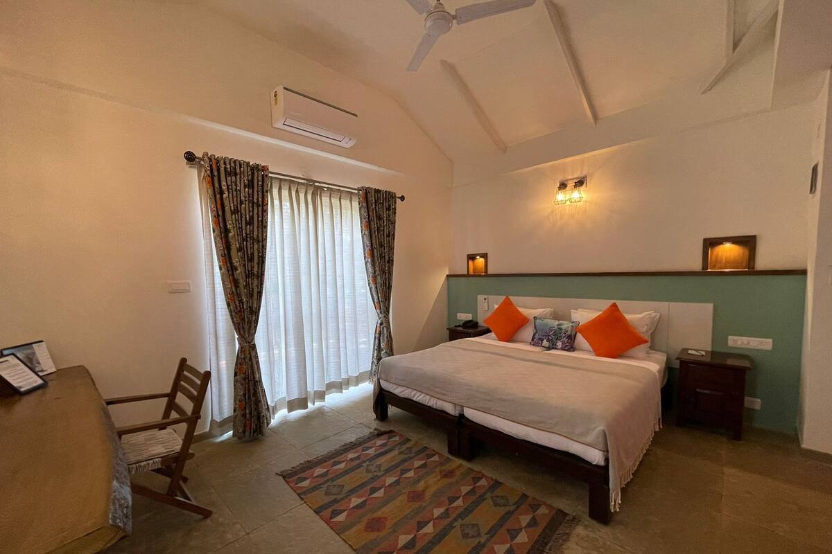 Habitación de hotel con cama, escritorio, silla, ventana con cortinas, unidad de aire acondicionado, ventilador de techo y alfombra estampada.