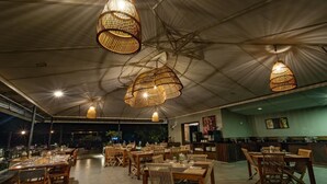 Un restaurante espacioso con techos altos y lámparas de mimbre colgantes. Las mesas están dispuestas con sillas de madera, y se observa una barra con el menú.