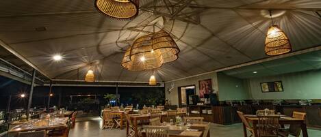 A spacious restaurant with high ceilings and hanging wicker lamps. Tables are set with wooden chairs, and a counter with a menu is visible.