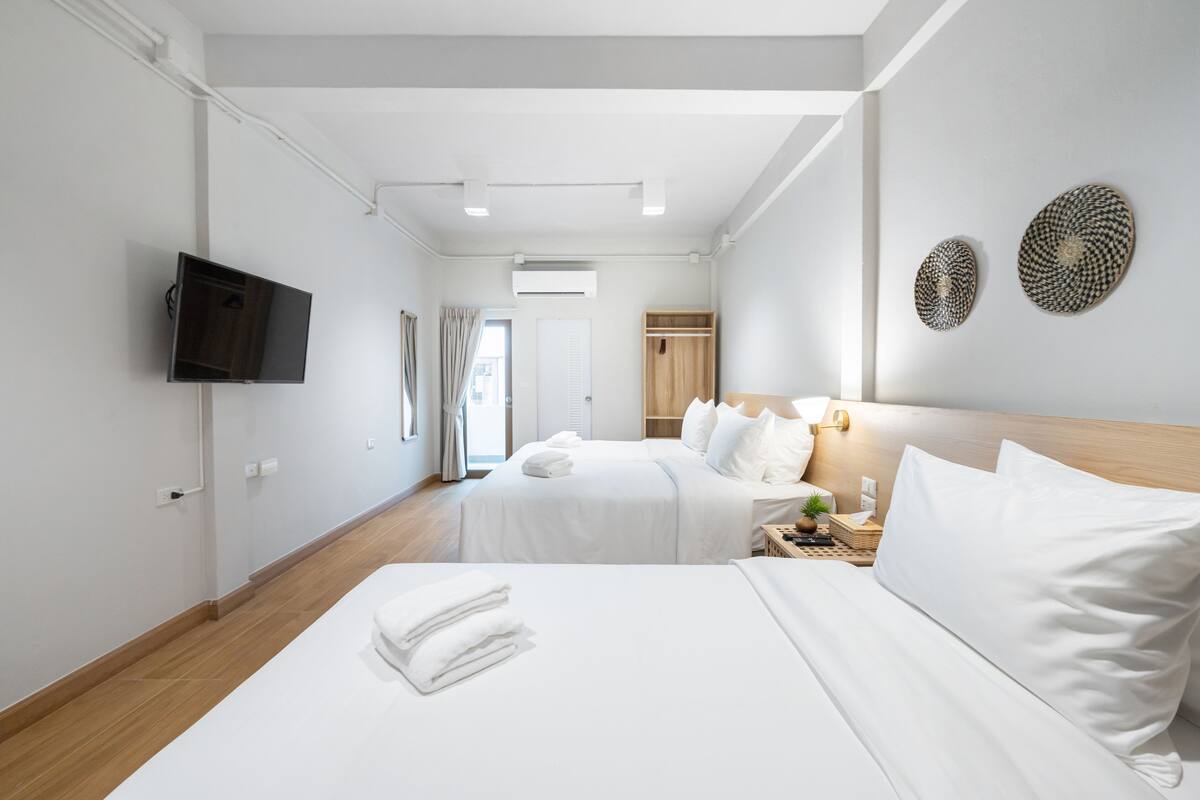 A hotel room with two beds, a TV mounted on the wall, a wooden wardrobe, and a bedside table with a lamp.