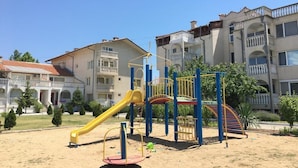 Un'area giochi con uno scivolo giallo e strutture per arrampicarsi, circondata da sabbia e edifici residenziali con balconi.